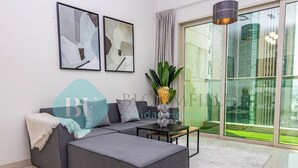 A modern living room with a grey sectional sofa, two abstract paintings, a potted plant, and a view of a green landscape through large glass doors. - Bloomfeilds Royal 1Br Skyscraper (Abu Dhabi)