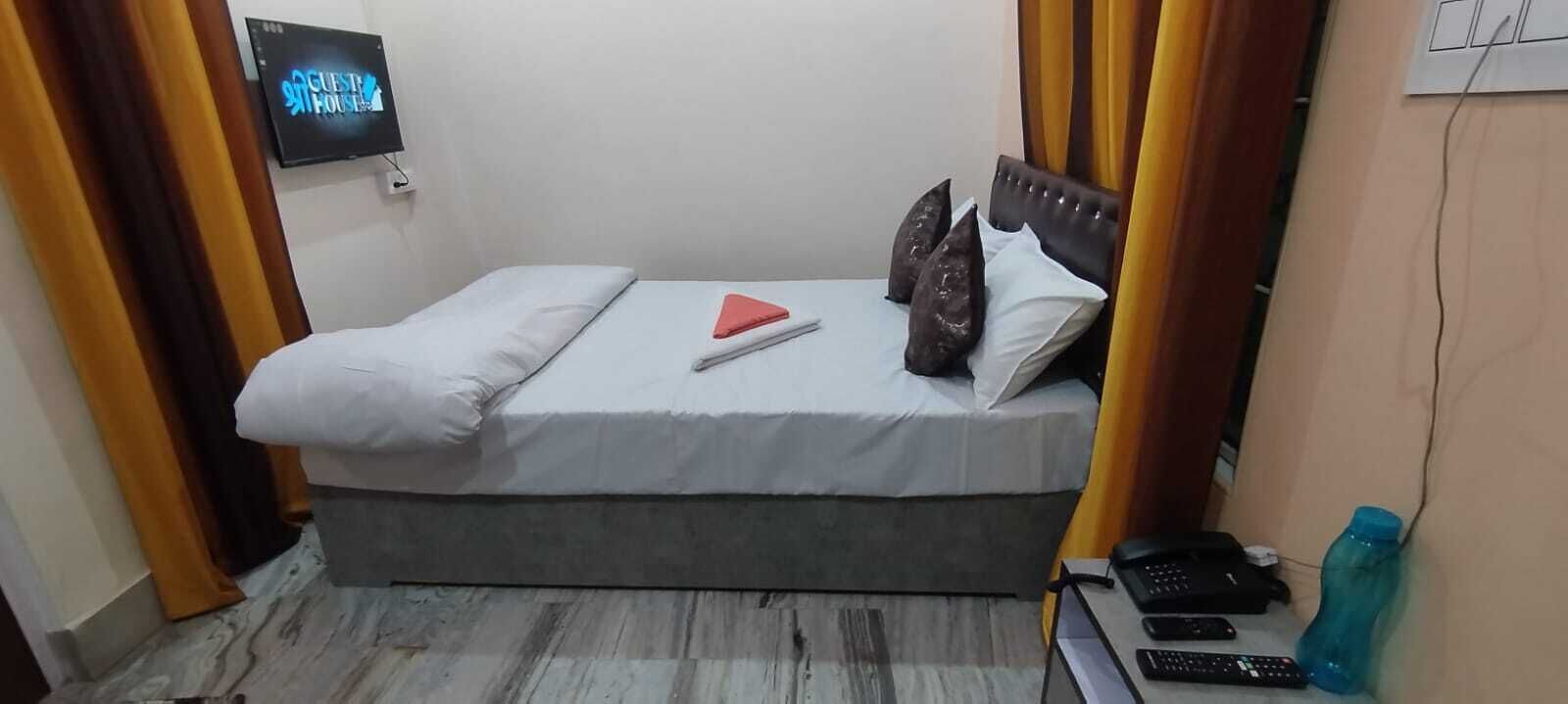 A single bed with a grey mattress, white and dark pillows, a red triangular object, and a black remote control on a wooden floor.