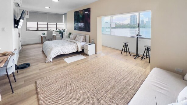 A modern bedroom with a large bed, a desk, and a view of the city through a large window. - La Rada 222 | Studio Apt in San Juan With Views (San Juan)