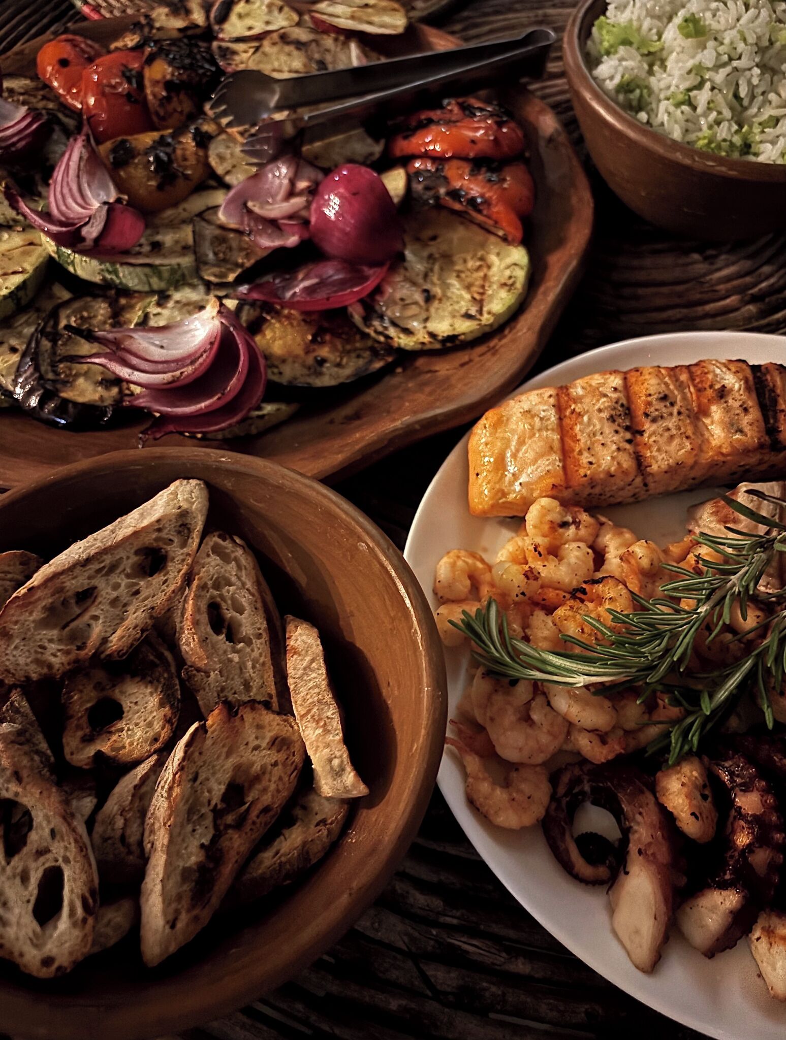A variety of dishes including grilled vegetables, shrimp, and grilled chicken, served on wooden plates.
