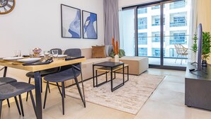 A modern living room with a sofa, dining table, and chairs. There is a large window with a view of buildings. - Bloomfields Stylish 1Br Binghatti Creek (Dubai)