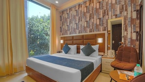Habitación de hotel con una cama grande, cabecera de madera, mesita de noche, una silla, una pequeña mesa y vistas a zonas verdes.