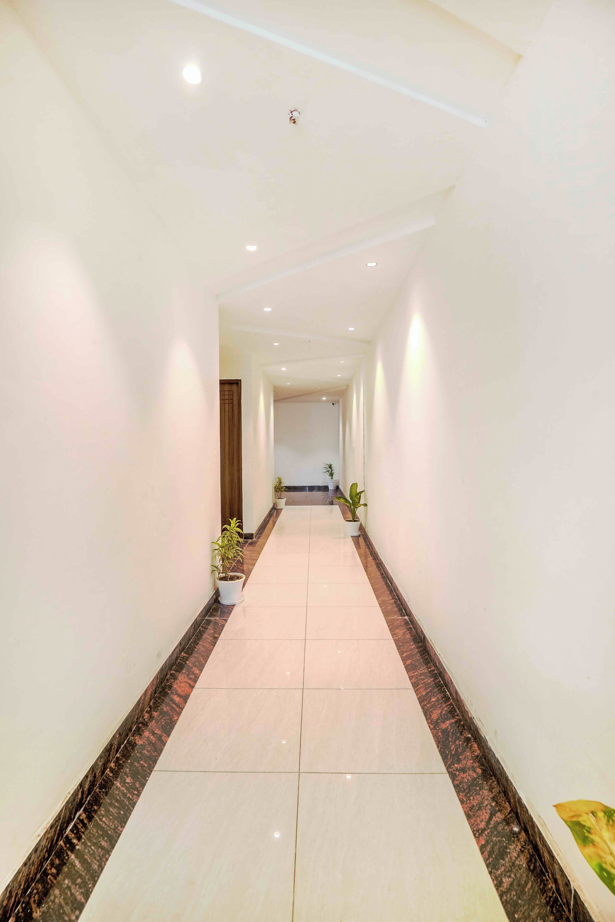 A long, narrow hallway with white walls and a marble floor. There are potted plants along the sides and a door at the end.