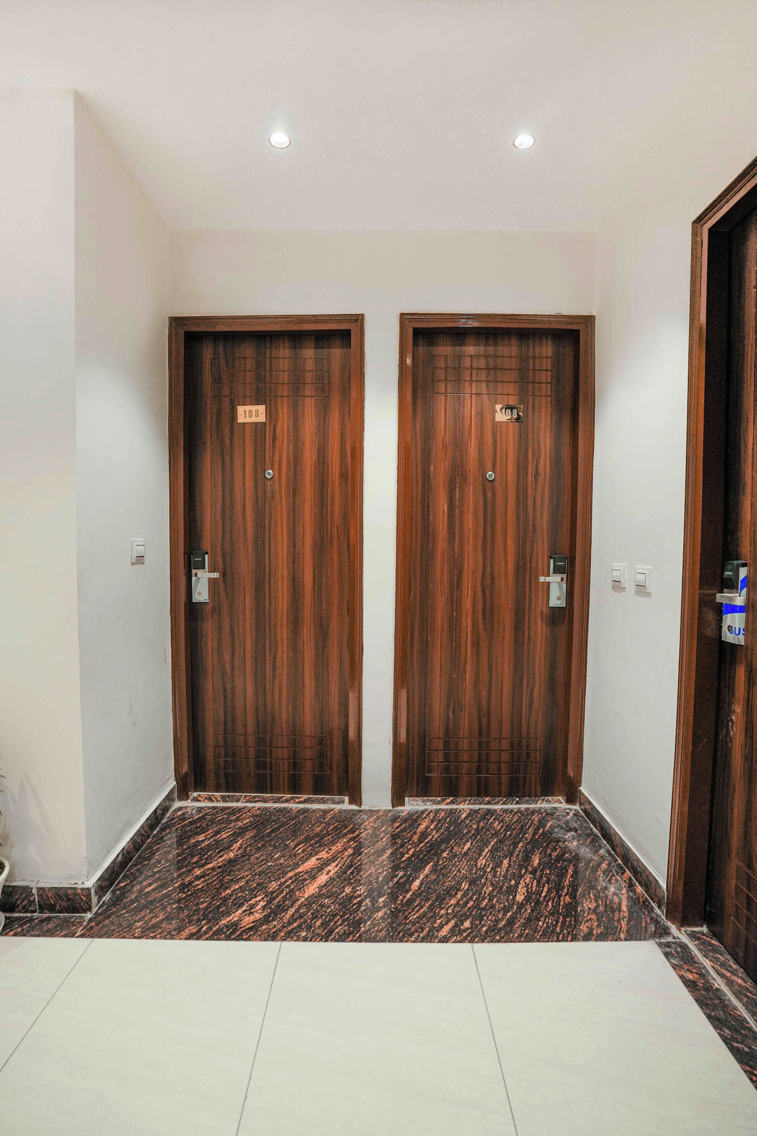Un pasillo con dos puertas de madera, cada una con una placa numerada, y un suelo de mármol.