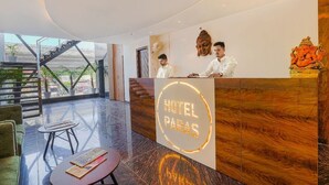 Zona de recepción de un hotel con diseño moderno, que cuenta con un mostrador de recepción con un cartel que dice “HOTEL PARAS”. Hay dos recepcionistas detrás del mostrador y una estatua de una divinidad es visible a la derecha. La zona de recepción tiene una pared de vidrio y vista al exterior.