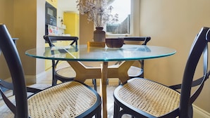 Una mesa redonda de vidrio con cuatro sillas negras, un cuenco de madera y un florero con flores secas.