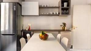 Une cuisine moderne avec un réfrigérateur en acier inoxydable, une table à manger avec des chaises et un panier de fruits. Des étagères au-dessus du comptoir servent à ranger les tasses et les verres.
