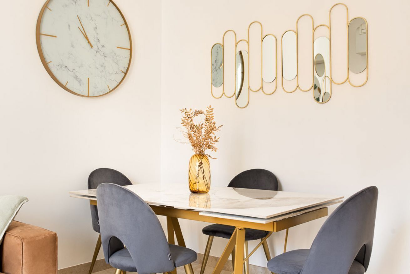 A dining area with a marble table, four chairs, a vase with dried flowers, a wall clock, and a series of mirrors.