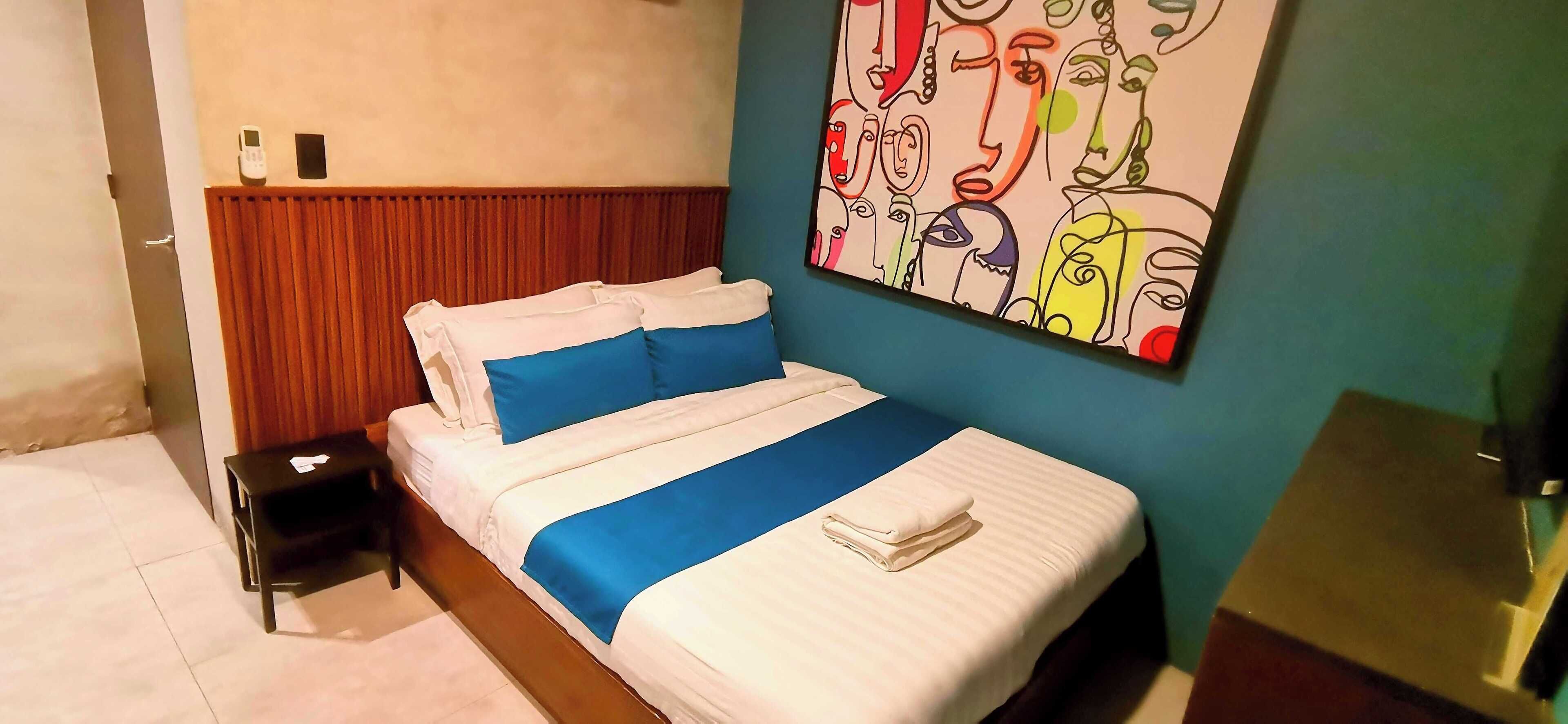 A hotel room with a bed, a nightstand, a painting on the wall, and a wooden headboard.