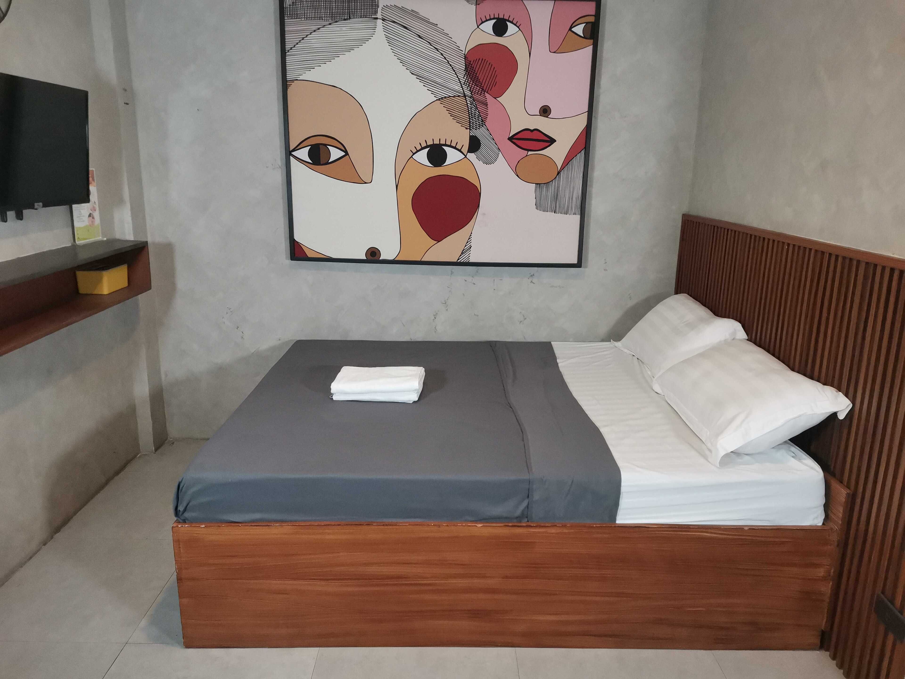 A bedroom with a wooden bed, grey and white bedding, a folded towel on the bed, and a painting on the wall.