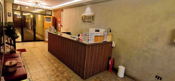 Zona de recepción de hotel con un mostrador de madera, un extintor, un cartel en la pared y una manguera contra incendios.