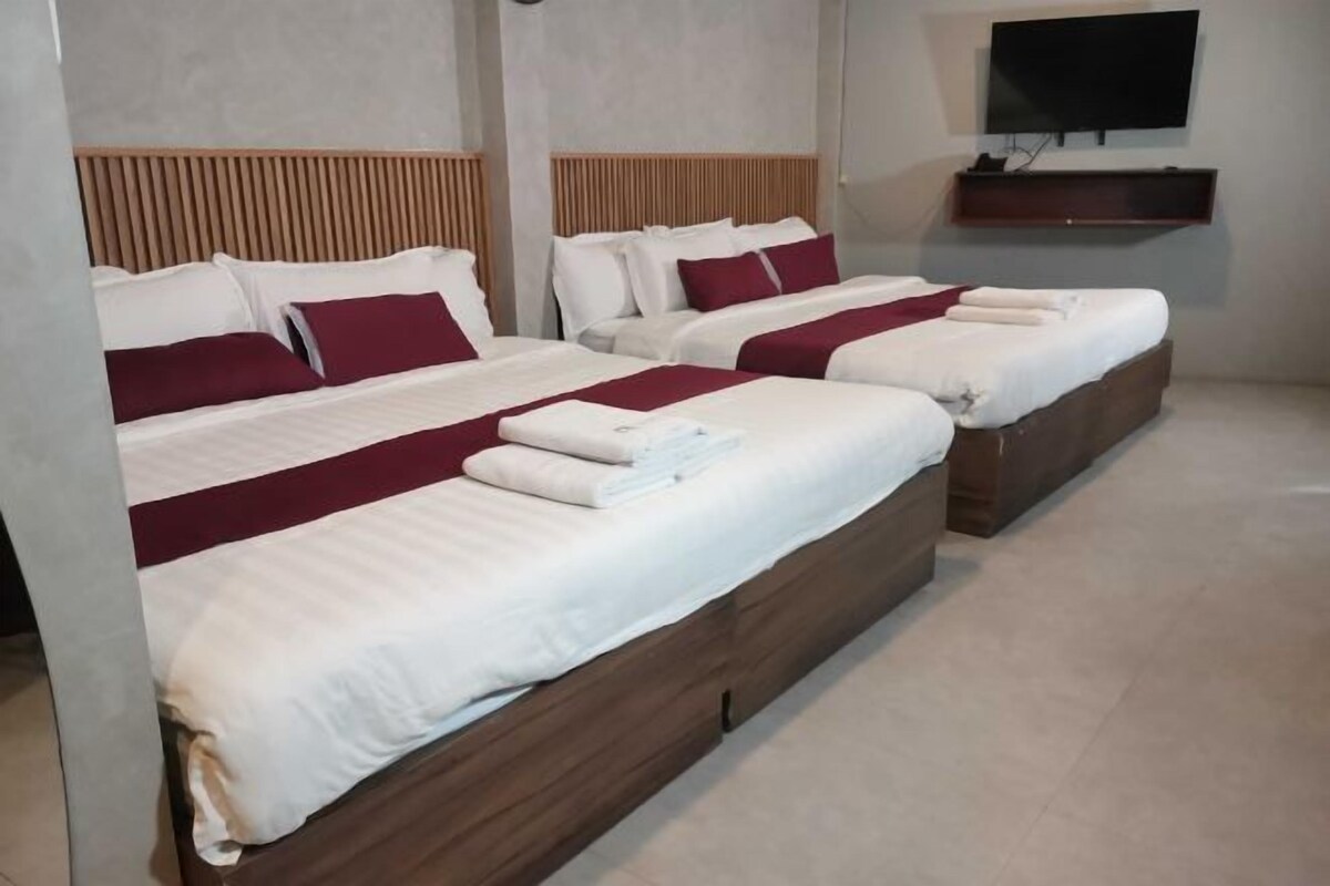 Habitación de hotel con tres camas, cada una con ropa blanca y detalles burdeos, cabeceros de madera, un televisor de pantalla plana montado en la pared y un estante pequeño debajo.