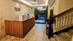 Un mostrador de recepción de hotel con un cartel encima, una escalera a la derecha y un suelo de baldosas.