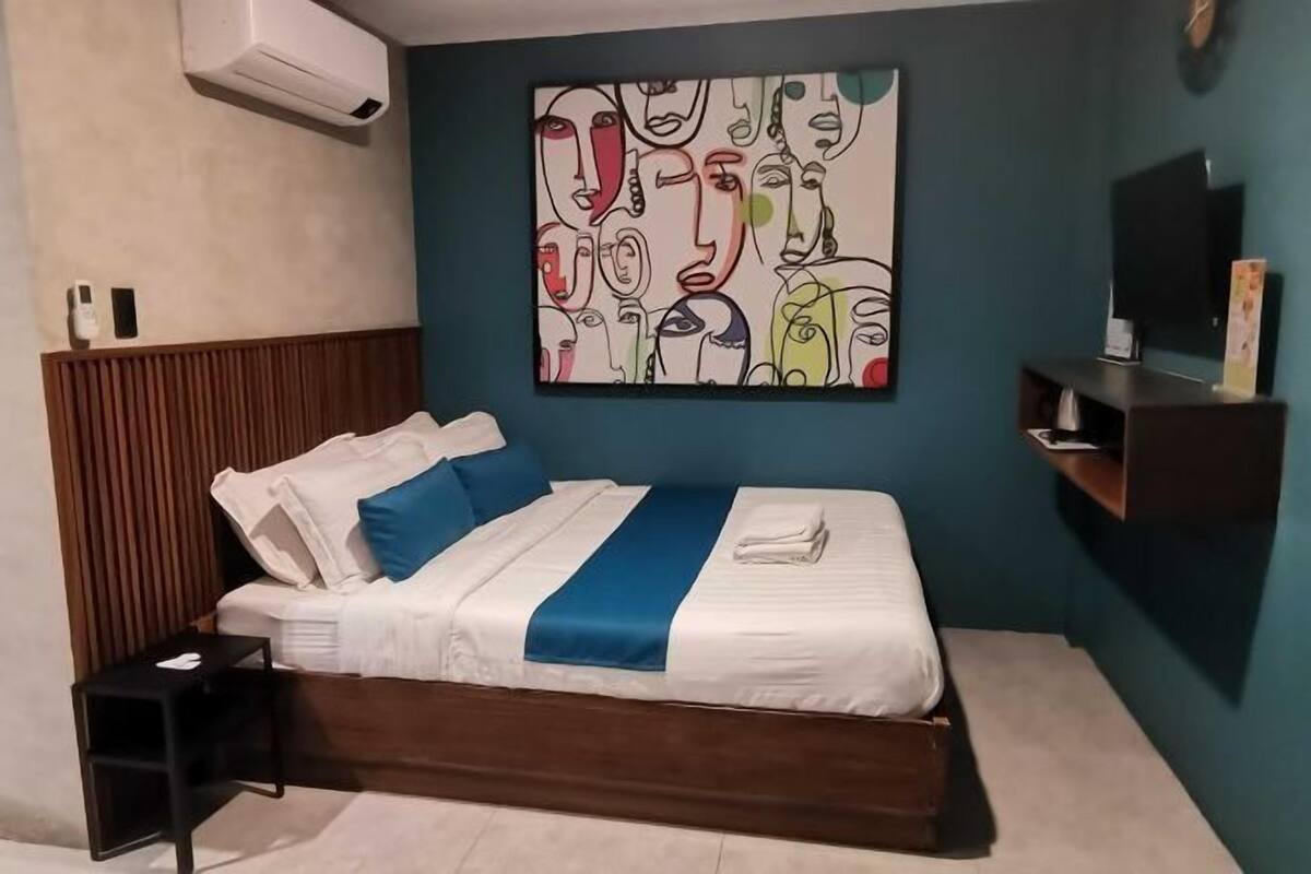 A hotel room with a bed, a wall-mounted air conditioner, a TV, and a shelf with items.