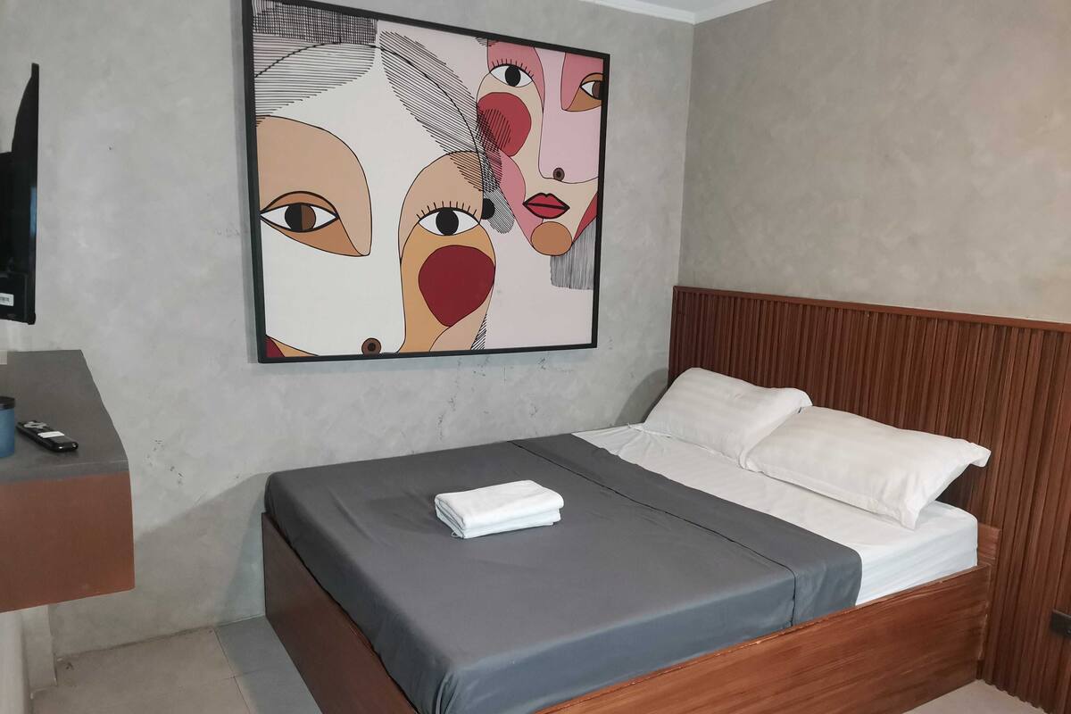 A hotel room with a wooden headboard, a grey bedspread, white pillows, and a framed abstract painting on the wall.