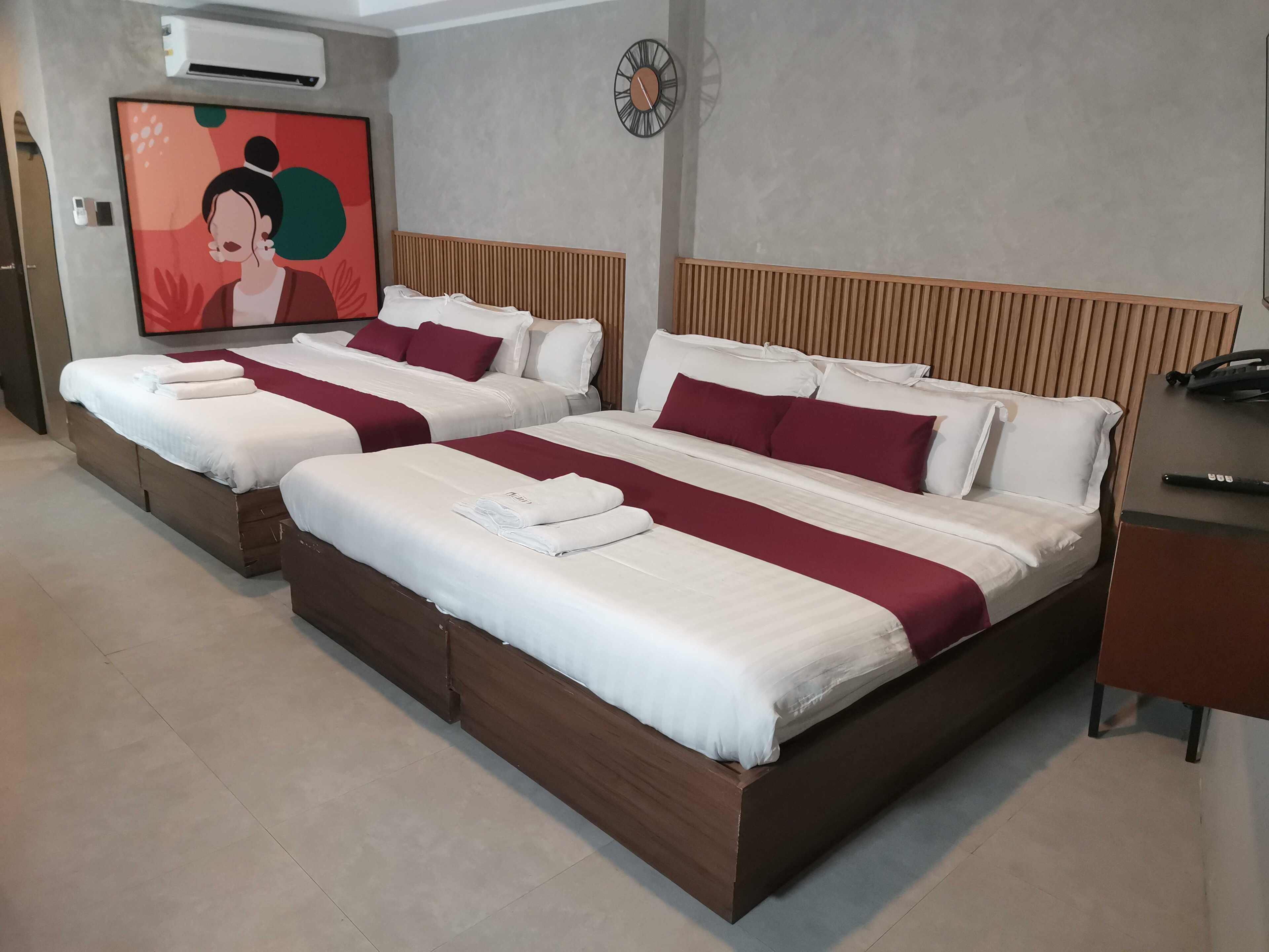A hotel room with two beds, a painting on the wall, a clock, and an air conditioning unit.