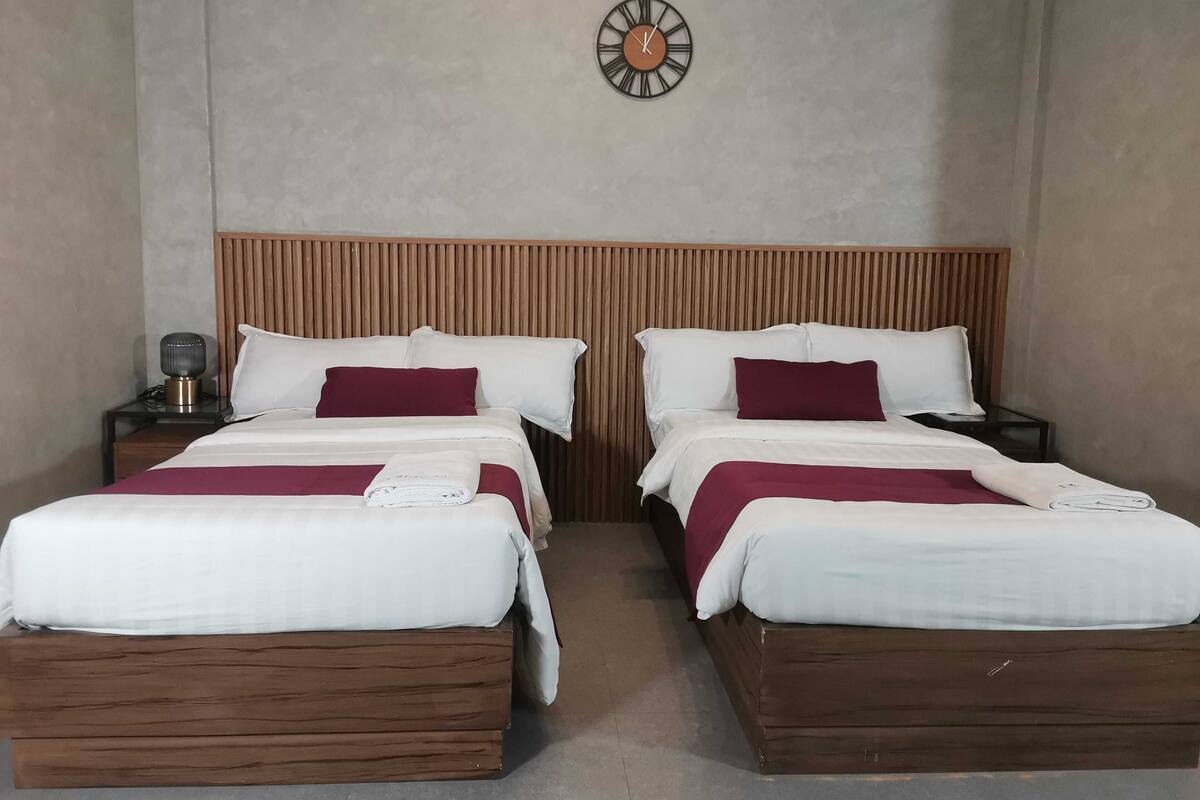 A hotel room with two beds, a wooden headboard, a clock on the wall, and a bedside table with a lamp.