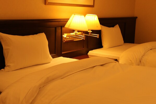 A hotel room with two beds, each with white linens and pillows, a nightstand with a lamp, and a wooden headboard. - Tsushima Grand Hotel (Tsushima)
