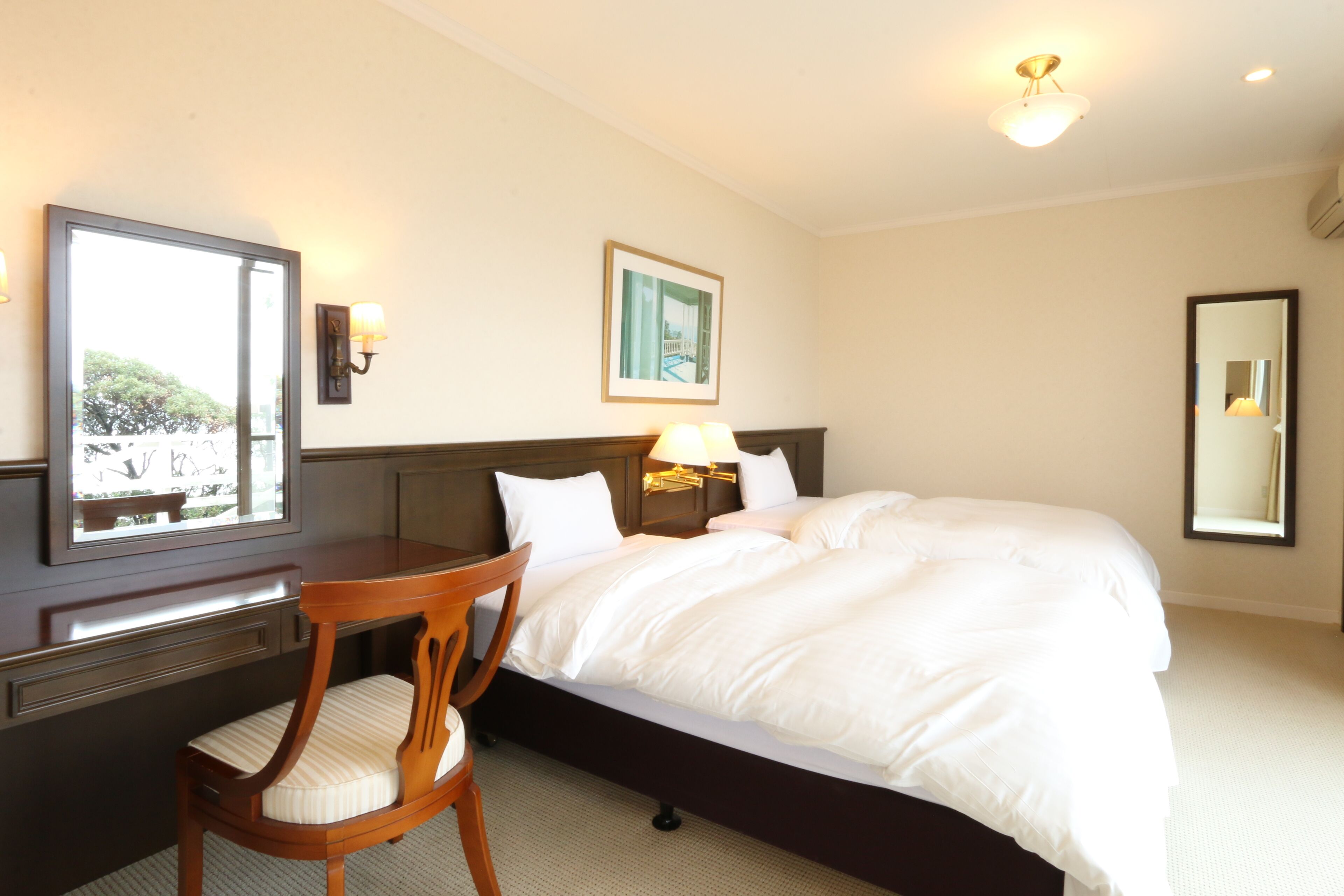 A hotel room with a large bed, a wooden chair, a desk, a desk lamp, a wall-mounted mirror, and a framed picture.