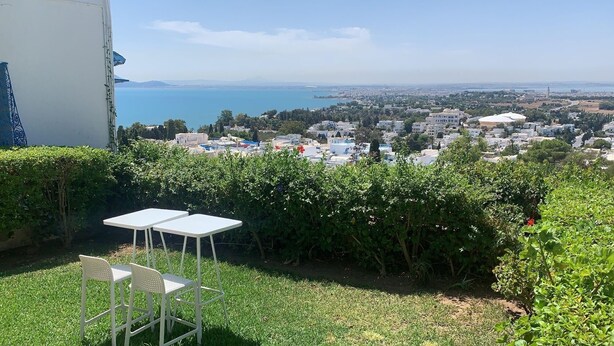 A terrace with two white tables and chairs, overlooking a coastal cityscape with buildings and a clear blue sea. - La Villa Des Oliviers (Site archéologique de Carthage)