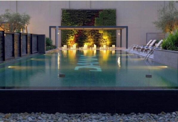 A modern indoor pool with a glass enclosure, a waterfall wall, and lounge chairs along the side.