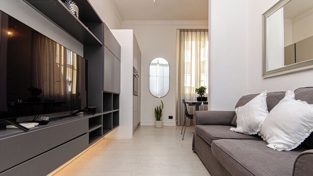 Una sala de estar moderna con un televisor de pantalla plana, un sofá gris con cojines blancos, una mesa de comedor con sillas y un espejo grande en la pared.