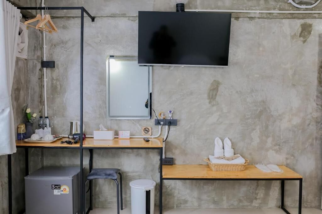 A small, functional room with a desk, a television mounted on the wall, a small heater, a trash bin, and a table with towels.