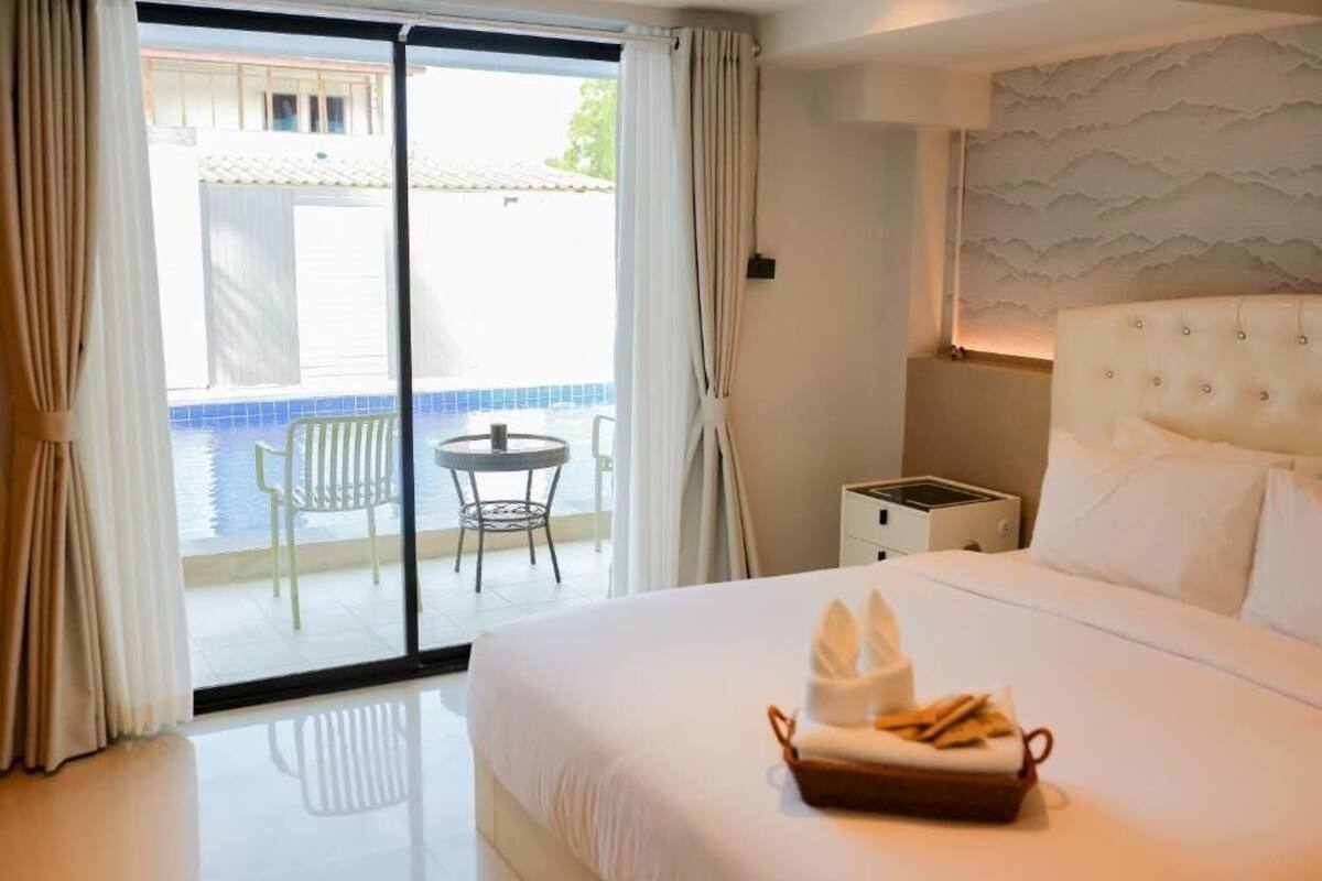 Ein Hotelzimmer mit einem großen Bett, einem kleinen Tisch, einem Sessel und Blick auf einen Pool durch eine Schiebetür.