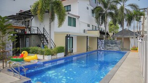 Una piscina con borde de azulejos, rodeada por un suelo de hormigón, palmeras y un edificio con balcón.