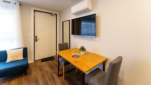 Una habitación con una mesa de comedor de madera, dos sillas, un sofá azul, una planta en maceta y un televisor montado.