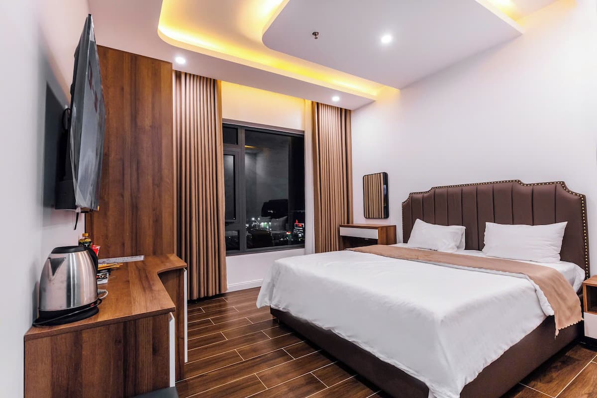 A modern hotel room with a large bed, a wooden desk, a flat-screen TV, and a view of the city at night.