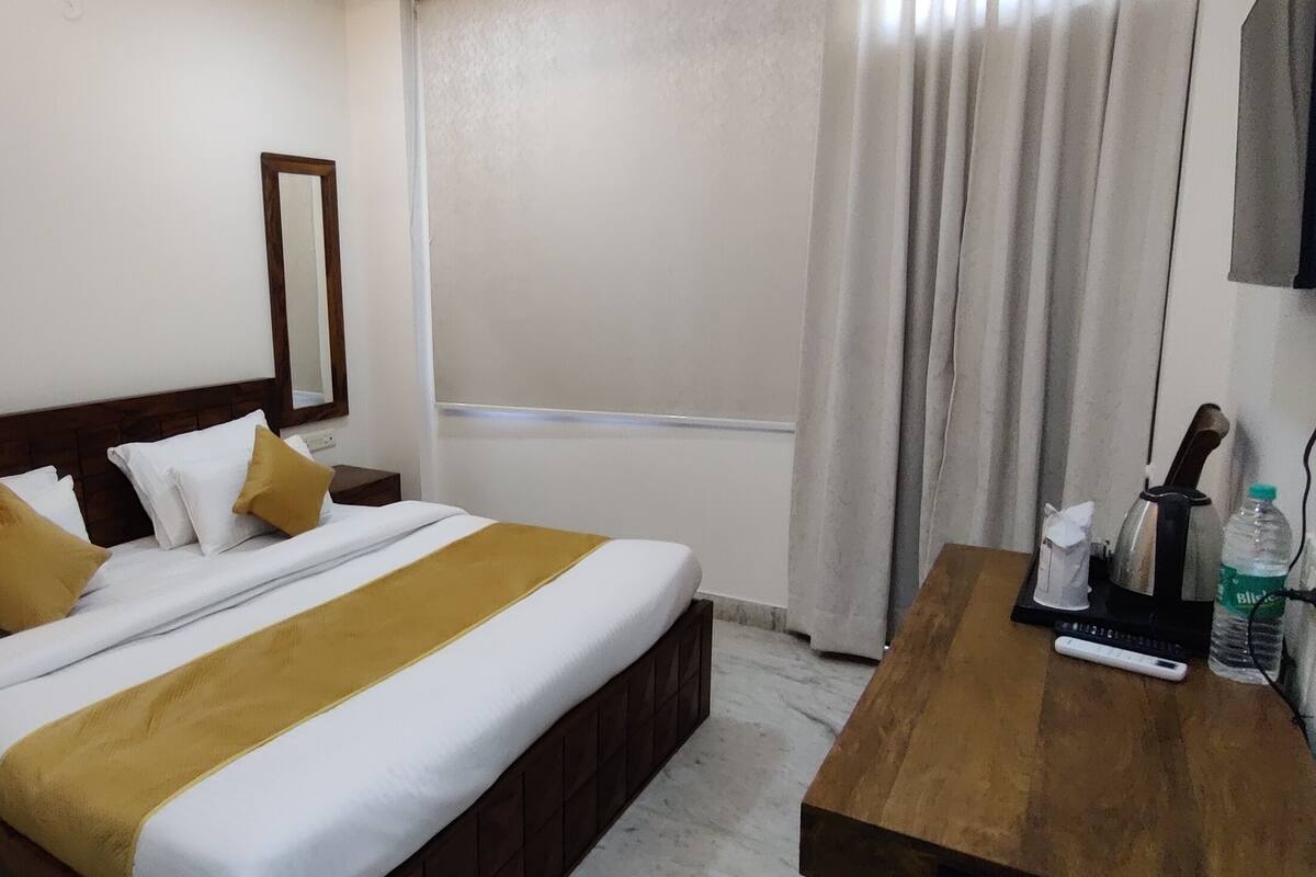 Ein Hotelzimmer mit einem Bett, einem Nachttisch aus Holz, einem Beistelltisch mit einer Flasche und einer Fernbedienung sowie einem an der Wand montierten Fernseher.