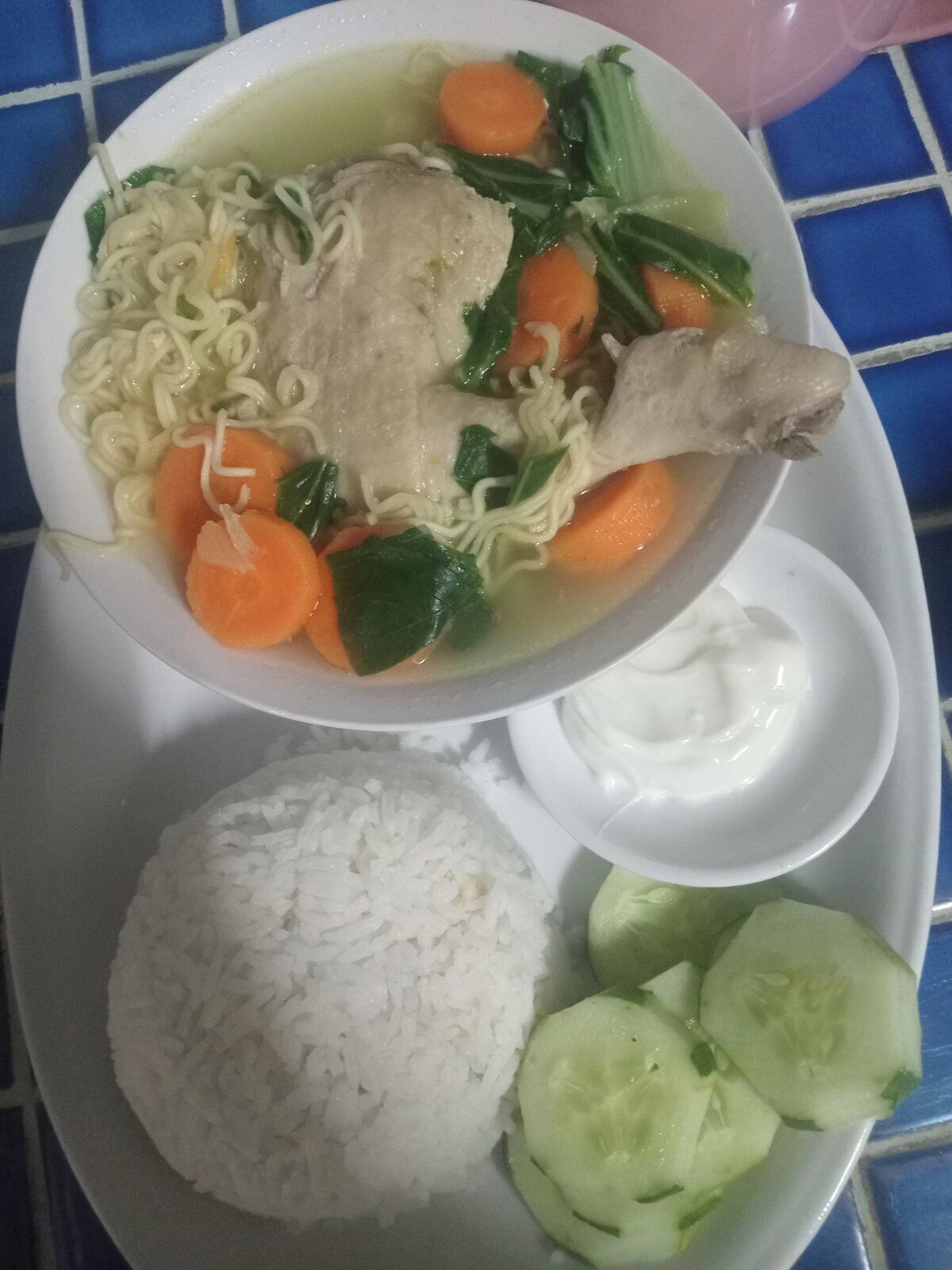 Une assiette avec un bol de soupe contenant des nouilles, du poulet, des carottes et des légumes verts, une portion de riz blanc et des concombres coupés en tranches.
