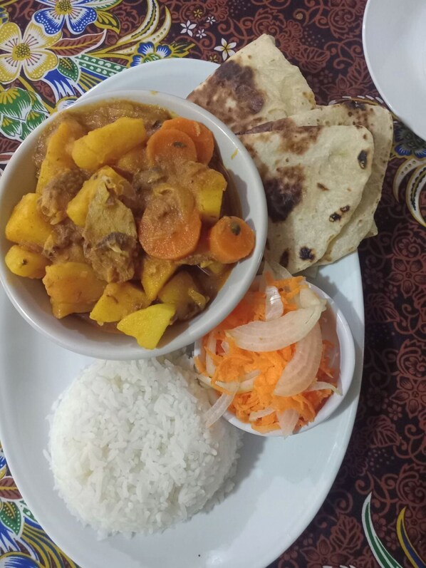 Une assiette de riz blanc, un bol de curry aux pommes de terre et aux carottes, un morceau de pain plat et un petit bol de carottes et d’oignons râpés.