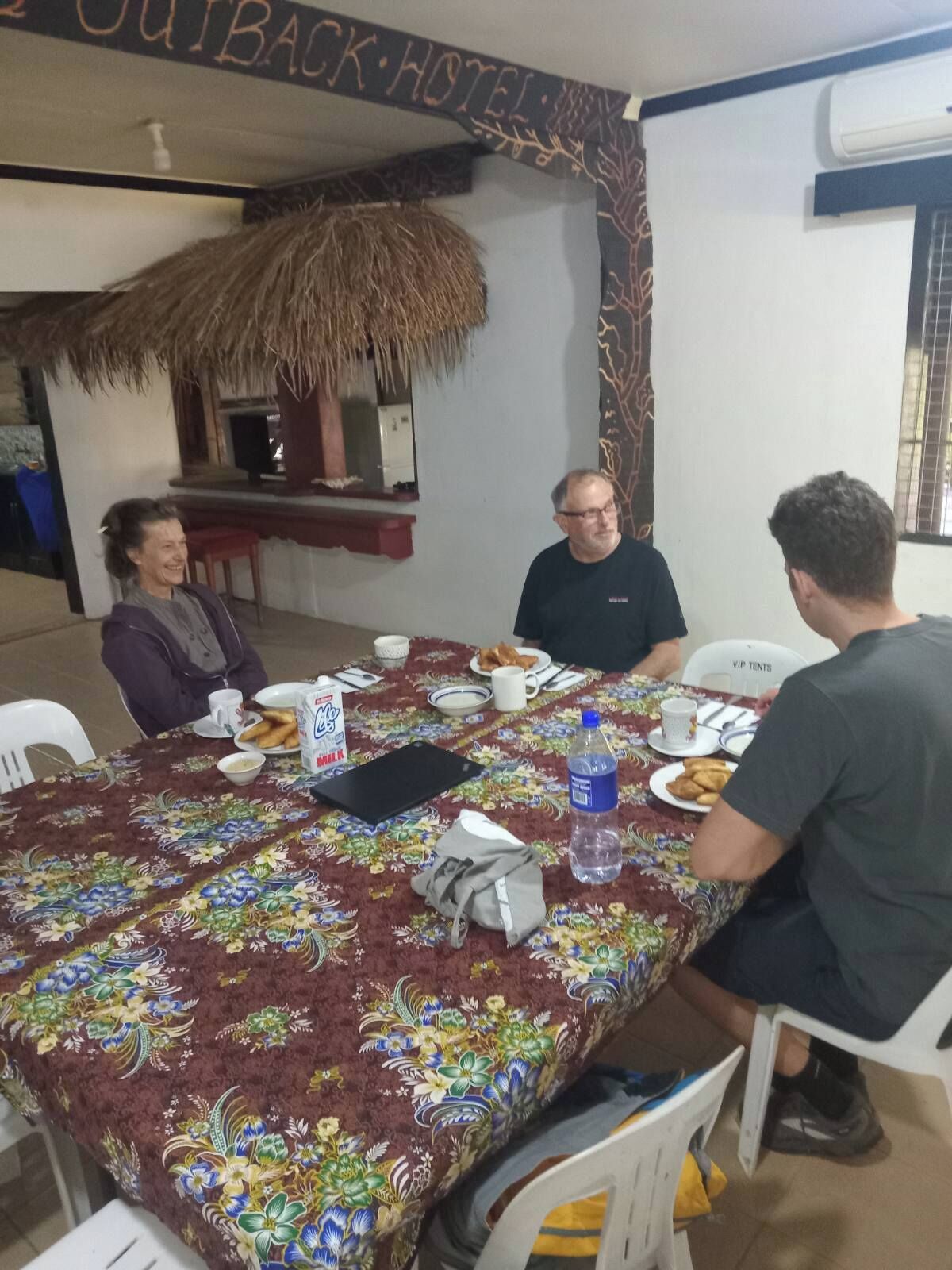 Un comedor con una mesa dispuesta para cuatro personas, una estructura de techo de paja y un letrero que dice 'OUTBACK HOTEL'.