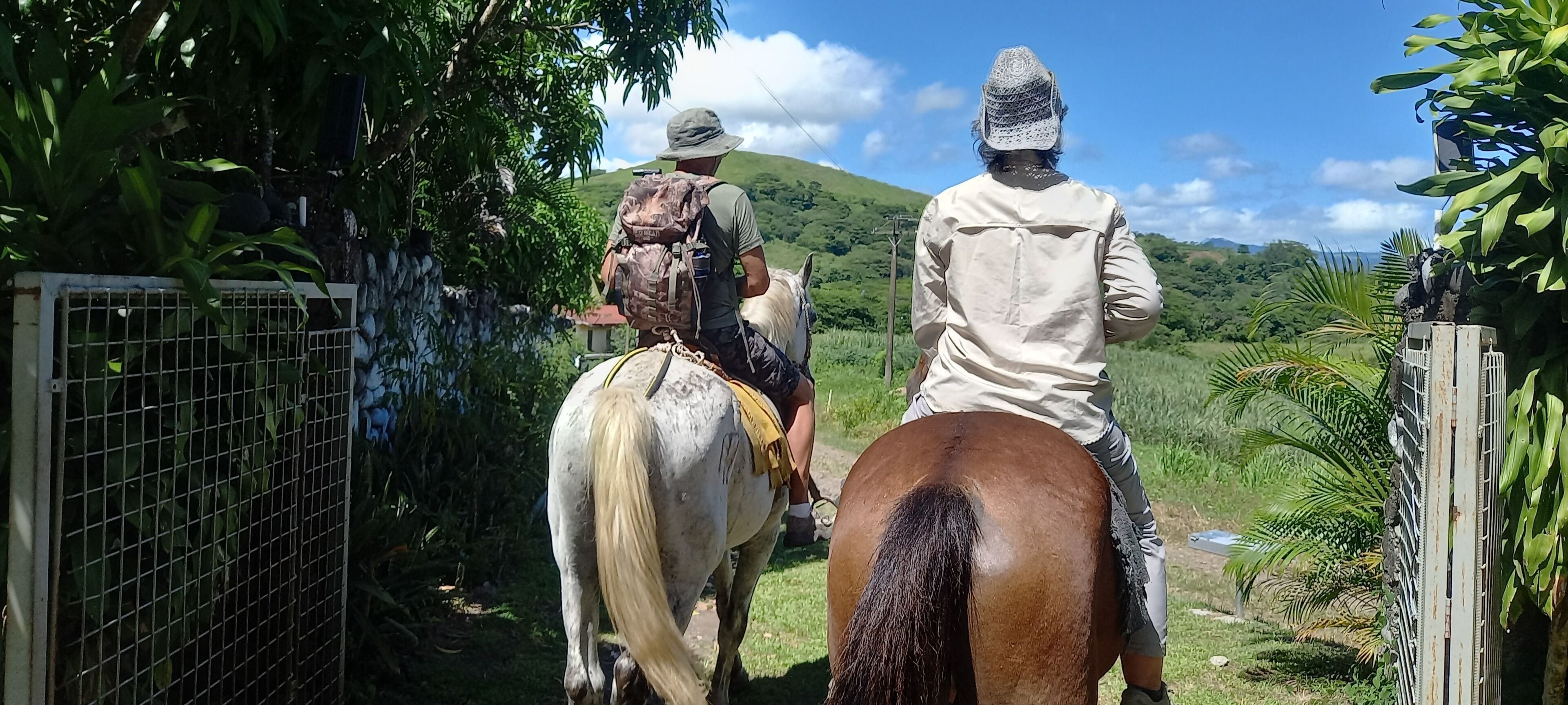 Dos personas montando a caballo por una puerta rodeada de follaje y colinas.