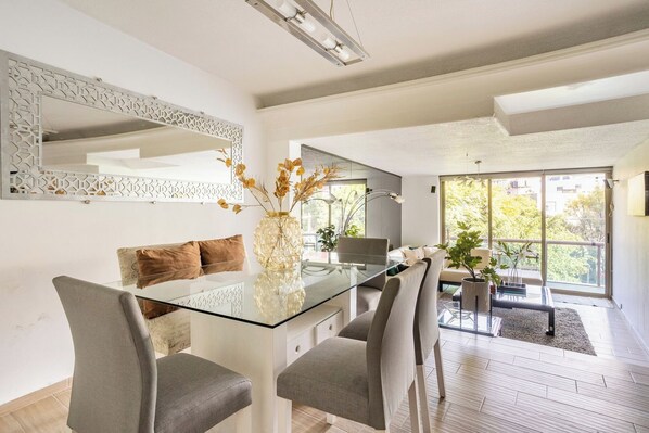 A modern dining area with a glass table, grey upholstered chairs, a decorative vase with dried flowers, and a large mirror on the wall.