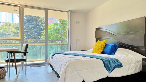 A modern bedroom with a large bed, a view of greenery through a sliding glass door, and a small table with a chair.