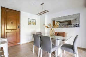 Un coin repas avec une table en verre, des chaises grises, un vase contenant des fleurs séchées et un tableau représentant un paysage urbain accroché au mur.