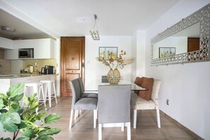 Een moderne keuken met witte kastjes, een magnetron en een koffieapparaat. Er is een eethoek met een tafel, stoelen en een vaas met gedroogde bloemen. Ook zichtbaar is een grote spiegel en een decoratief wandpaneel.