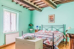 A room with a bed, a radiator, a window with curtains, a small table with a lamp, and a picture on the wall.