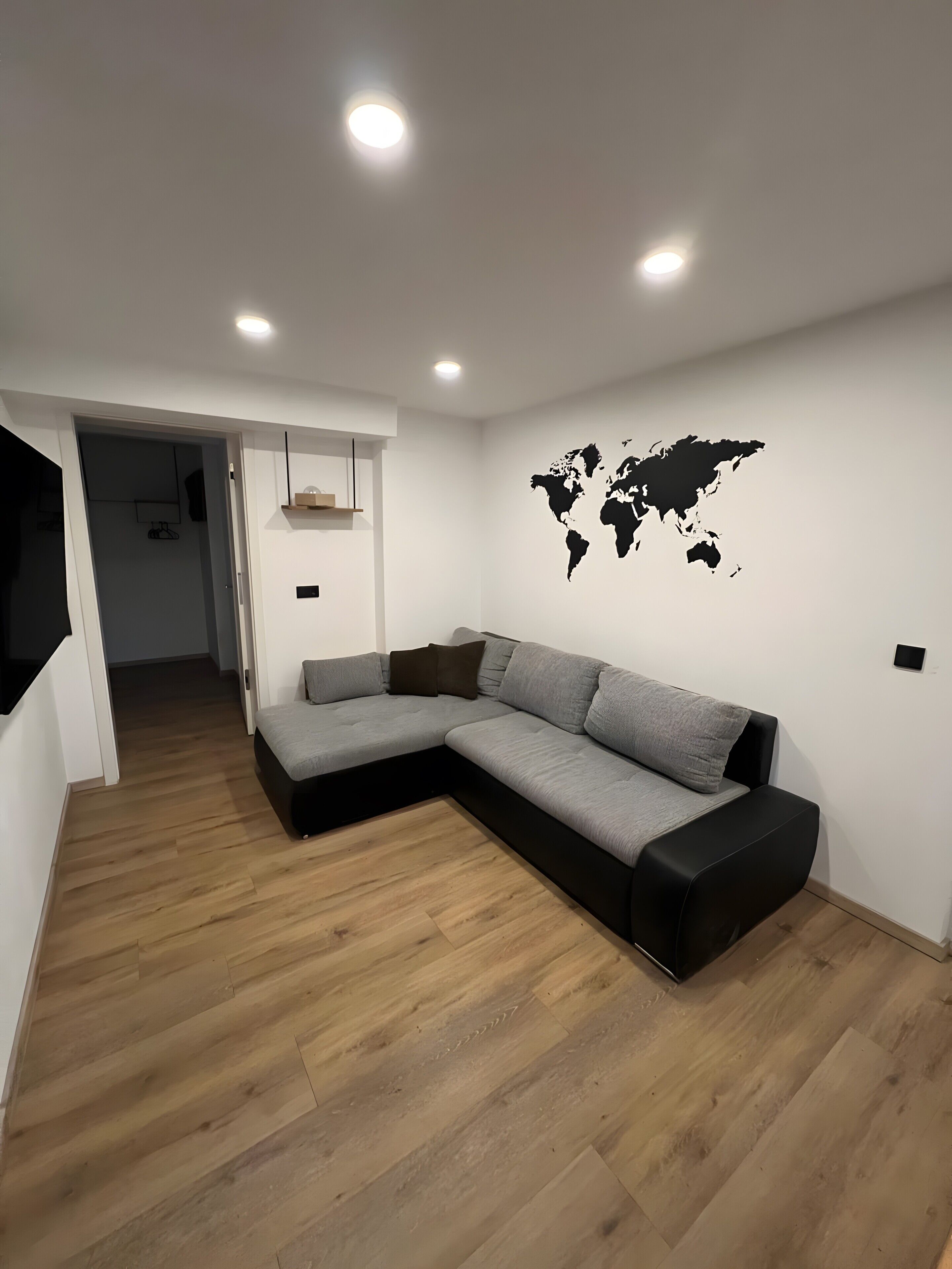 Un salon moderne avec un canapé d’angle, un plancher en bois, des spots encastrés au plafond, un grand autocollant représentant une carte du monde sur un mur, et une porte donnant sur une autre pièce.