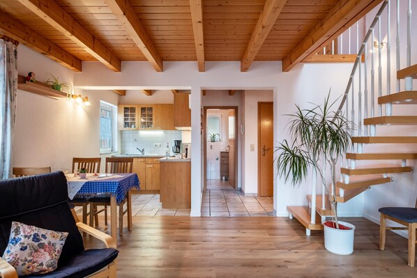 A spacious living area with a wooden staircase, a dining area with a blue tablecloth, and a kitchen in the background.