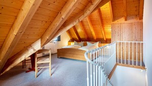 Een slaapkamer met schuine wanden, houten balken, een bed, een stoel en een witte leuning.
