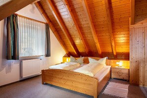 Une chambre avec un plafond en pente, un lit en bois, une table de chevet, une fenêtre avec des rideaux et un radiateur.