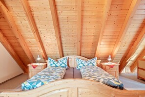 Quarto no sótão com teto inclinado de madeira, cama com roupa de cama estampada, abajures e uma pequena mesa de cabeceira de madeira.