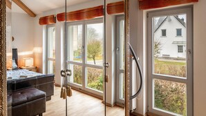 Ein Zimmer mit Bett, Schreibtisch, Stuhl, einem großen Spiegel und Blick auf ein Haus durch die Fenster.