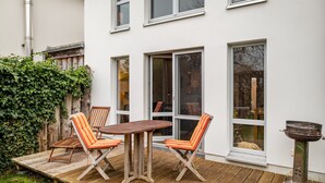 Eine Terrasse mit einem Holztisch und -stühlen, einem Grill und einem weißen Gebäude mit großen Fenstern.