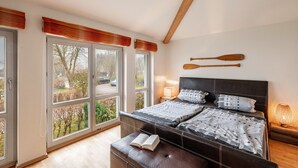 Ein Schlafzimmer mit großem Fenster, einem Bett mit Kopfteil, einem Nachttisch mit Lampe und einem Buch auf der Bank.