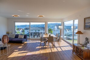 Ampla sala de estar com uma grande mesa de jantar, sofá e vista para um lago e prédios.